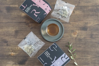 A cup of tea sits on a gray saucer, surrounded by two distinctively designed tea boxes and two individually wrapped tea bags, all placed on a wooden surface. An olive branch is also present, adding a natural element to the arrangement.