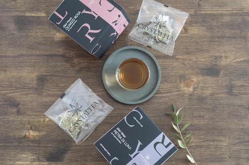 A cup of tea sits on a gray saucer, surrounded by two distinctively designed tea boxes and two individually wrapped tea bags, all placed on a wooden surface. An olive branch is also present, adding a natural element to the arrangement.
