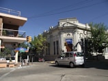 A street scene featuring an ornate white building marked with 'Banco Republica' and a smaller commercial building with a balcony. A silver car is driving on the street. There are traffic lights and trees around the area, along with an umbrella and some outdoor seating near the commercial building.
