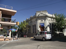 A street scene featuring an ornate white building marked with 'Banco Republica' and a smaller commercial building with a balcony. A silver car is driving on the street. There are traffic lights and trees around the area, along with an umbrella and some outdoor seating near the commercial building.