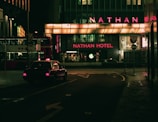 A dimly lit urban street scene at night featuring a taxi driving past a building with prominent neon lighting. The signage highlights 'Nathan Hotel' in bright red, while a 'Starbucks' logo is visible nearby. A double-decker bus is parked on the side, partially visible with Chinese characters.