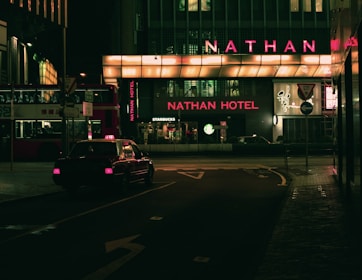 A dimly lit urban street scene at night featuring a taxi driving past a building with prominent neon lighting. The signage highlights 'Nathan Hotel' in bright red, while a 'Starbucks' logo is visible nearby. A double-decker bus is parked on the side, partially visible with Chinese characters.