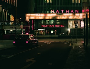 A dimly lit urban street scene at night featuring a taxi driving past a building with prominent neon lighting. The signage highlights 'Nathan Hotel' in bright red, while a 'Starbucks' logo is visible nearby. A double-decker bus is parked on the side, partially visible with Chinese characters.
