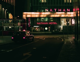 A dimly lit urban street scene at night featuring a taxi driving past a building with prominent neon lighting. The signage highlights 'Nathan Hotel' in bright red, while a 'Starbucks' logo is visible nearby. A double-decker bus is parked on the side, partially visible with Chinese characters.