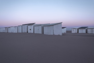 A row of white beach cabins stands on a sandy beach under a serene sky. The cabins are arranged closely, with simple structures and visible doors. The sky exhibits a gradient of pastel colors, transitioning from light blue to pink, suggesting either early morning or evening.