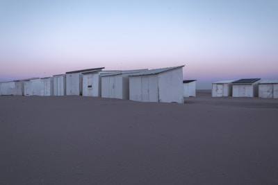 A row of white beach cabins stands on a sandy beach under a serene sky. The cabins are arranged closely, with simple structures and visible doors. The sky exhibits a gradient of pastel colors, transitioning from light blue to pink, suggesting either early morning or evening.