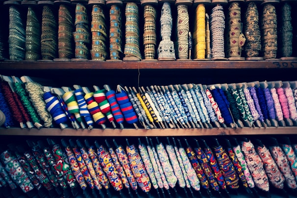 A colorful display of various spools of thread and ribbons arranged on shelves. The spools come in multiple patterns, textures, and colors, showcasing a diverse range of embroidery and fabric art. Intricate designs and a variety of textiles are visible, highlighting the rich craft of fabric embellishments.