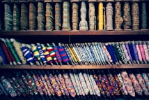 A colorful display of various spools of thread and ribbons arranged on shelves. The spools come in multiple patterns, textures, and colors, showcasing a diverse range of embroidery and fabric art. Intricate designs and a variety of textiles are visible, highlighting the rich craft of fabric embellishments.