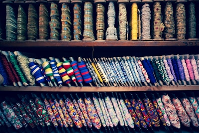 A colorful display of various spools of thread and ribbons arranged on shelves. The spools come in multiple patterns, textures, and colors, showcasing a diverse range of embroidery and fabric art. Intricate designs and a variety of textiles are visible, highlighting the rich craft of fabric embellishments.