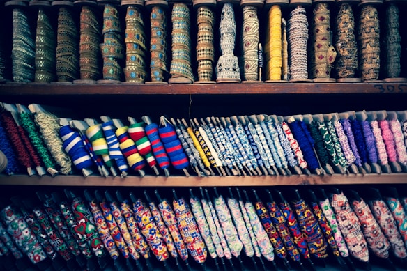 A colorful display of various spools of thread and ribbons arranged on shelves. The spools come in multiple patterns, textures, and colors, showcasing a diverse range of embroidery and fabric art. Intricate designs and a variety of textiles are visible, highlighting the rich craft of fabric embellishments.
