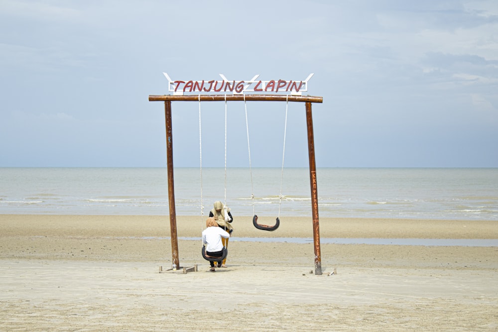 man in white t-shirt and black pants sitting on brown wooden swing chair on beach