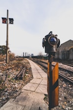 A railway track scene with a focus on a vintage-style signal light and a small signaling device in the foreground. The background shows multiple railway tracks, a pathway, and distant buildings under a clear sky. A digital clock display is visible on a pole, showing 12:18.