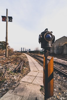A railway track scene with a focus on a vintage-style signal light and a small signaling device in the foreground. The background shows multiple railway tracks, a pathway, and distant buildings under a clear sky. A digital clock display is visible on a pole, showing 12:18.