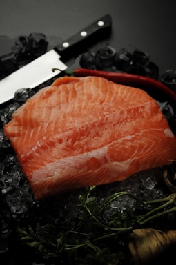 A raw salmon fillet is placed on top of crushed ice, accompanied by a large chef's knife with a black handle. A red chili pepper and some fresh green herbs are also present, suggesting preparation for cooking.