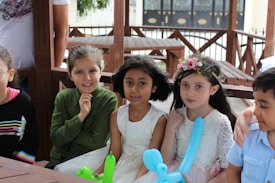 A group of children sitting together on wooden benches. The two girls in the center are wearing white dresses, one of whom has a flower crown, while the girl on the left is wearing a green top. They are holding balloon animals and appear to be enjoying themselves. A boy on the right is wearing a light blue shirt.