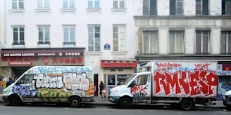 Two vans are parked on a street, covered in colorful graffiti. The background features an old building with several windows and a couple of storefronts, including a supermarket with signs in both English and Chinese.