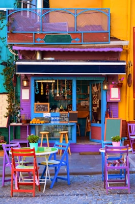A vibrant, colorful outdoor cafe with multicolored chairs and tables arranged on a cobblestone street. The facade of the cafe is painted in bright shades, including purple, blue, and yellow. Hanging decorations and strings of beads adorn the front of the cafe. A small chalkboard sign reads 'Coffee To Go' and there are various items on the counter such as water bottles, oranges, and small ornaments. Potted plants add a touch of greenery to the scene.