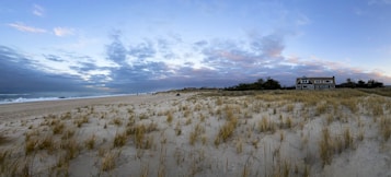 A coastal landscape featuring a sandy beach with scattered grasses leading up to a distant shoreline. The sky is filled with a mix of blue and gray clouds, suggesting early morning or late evening. On the right side, a large beach house surrounded by shrubs and trees adds a sheltered, inviting element to the scene.