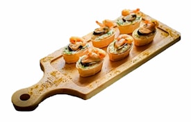 A wooden serving board holds six pastry cups filled with a creamy mixture, garnished with shrimp and mushrooms. The pastry cups have a light golden-brown crust, and the toppings are arranged attractively.