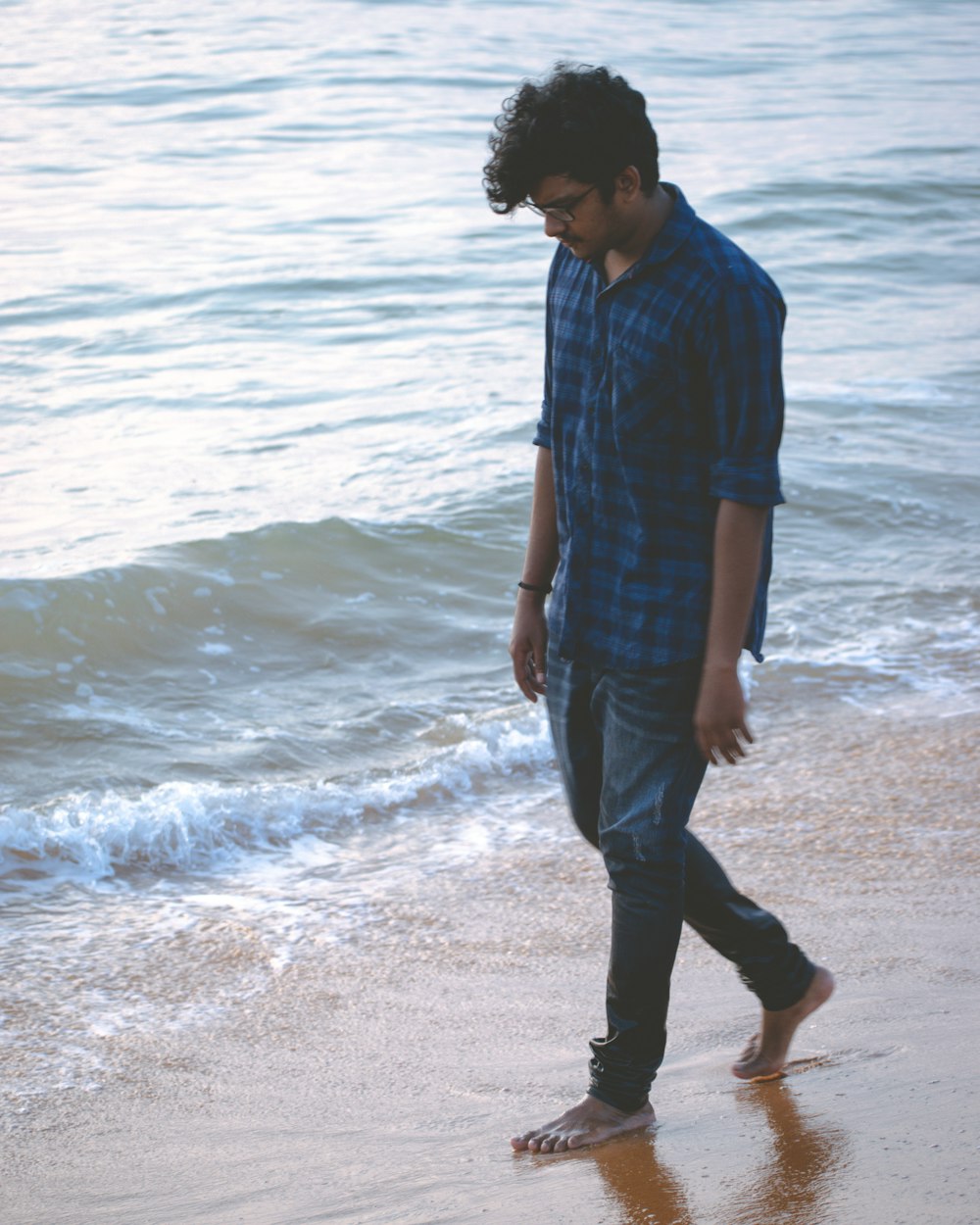 Hombre con camisa de vestir a cuadros azul y negro y pantalones negros de pie en la orilla del mar durante el día