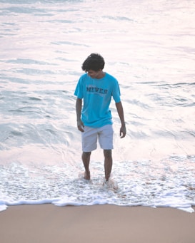 A person is standing in shallow ocean waves near the shoreline, wearing a light blue shirt with text and light-colored shorts. The ocean water looks calm, and light reflects off its surface, creating a serene and peaceful atmosphere.