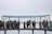 A group of people is gathered on a metal observation platform suspended in the air. The platform has a roof with a blue tint and is surrounded by a metal grid structure for safety. The sky appears overcast, giving the scene a slightly gloomy or neutral tone. Individuals are looking through the grid, possibly observing a view or landscape.