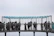 A group of people is gathered on a metal observation platform suspended in the air. The platform has a roof with a blue tint and is surrounded by a metal grid structure for safety. The sky appears overcast, giving the scene a slightly gloomy or neutral tone. Individuals are looking through the grid, possibly observing a view or landscape.