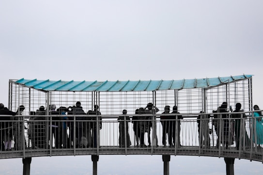 A group of people is gathered on a metal observation platform suspended in the air. The platform has a roof with a blue tint and is surrounded by a metal grid structure for safety. The sky appears overcast, giving the scene a slightly gloomy or neutral tone. Individuals are looking through the grid, possibly observing a view or landscape.
