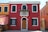 A colorful building facade with a vibrant red exterior, accented by white window frames and a wooden door. Green shutters adorn the windows, and there is a small balcony with a flower arrangement. Adjacent buildings are painted in bold orange and yellow hues.