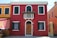 A colorful building facade with a vibrant red exterior, accented by white window frames and a wooden door. Green shutters adorn the windows, and there is a small balcony with a flower arrangement. Adjacent buildings are painted in bold orange and yellow hues.