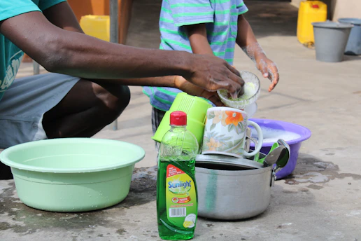 Close-up of a hand washing dishes with natural orange-colored dishwash liquid surrounded by fresh herbs.