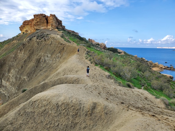 A hiker walks along a narrow trail atop a rugged hillside with a view of the ocean in the background. The path cuts through green vegetation and moves towards a rocky outcrop. The sky is partly cloudy, adding a dynamic element to the landscape.