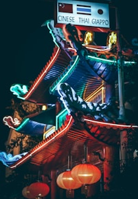 A vibrant, neon-lit structure featuring traditional Asian architectural elements. Brightly colored signs with flags and words like 'CINESE,' 'THAI,' and 'GIAPPO' are visible above. Red lanterns hang below, casting a warm glow.