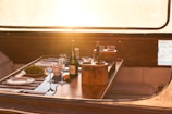 A wooden tabletop on a boat is set for a meal with plates, wine glasses, and a bottle of champagne. A bowl of grapes and an orange are placed next to a wooden caddy with more glasses and another bottle. Sunlight streams through a window, creating a warm, inviting atmosphere.