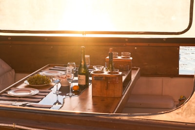 A wooden tabletop on a boat is set for a meal with plates, wine glasses, and a bottle of champagne. A bowl of grapes and an orange are placed next to a wooden caddy with more glasses and another bottle. Sunlight streams through a window, creating a warm, inviting atmosphere.