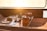 A wooden tabletop on a boat is set for a meal with plates, wine glasses, and a bottle of champagne. A bowl of grapes and an orange are placed next to a wooden caddy with more glasses and another bottle. Sunlight streams through a window, creating a warm, inviting atmosphere.