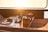 A wooden tabletop on a boat is set for a meal with plates, wine glasses, and a bottle of champagne. A bowl of grapes and an orange are placed next to a wooden caddy with more glasses and another bottle. Sunlight streams through a window, creating a warm, inviting atmosphere.