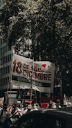 A group of people participate in a protest or gathering on a city street, holding a large banner with bold text in Spanish and various signs. The scene is shaded by large tree branches, and some participants are wearing masks, possibly indicating a recent time period.