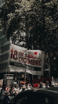 A group of people participate in a protest or gathering on a city street, holding a large banner with bold text in Spanish and various signs. The scene is shaded by large tree branches, and some participants are wearing masks, possibly indicating a recent time period.