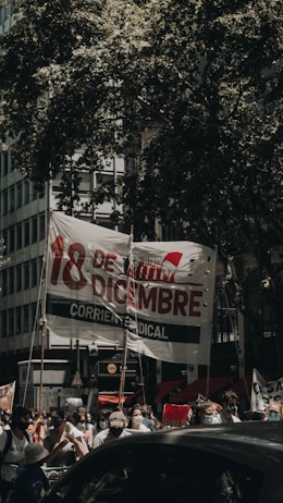 A group of people participate in a protest or gathering on a city street, holding a large banner with bold text in Spanish and various signs. The scene is shaded by large tree branches, and some participants are wearing masks, possibly indicating a recent time period.