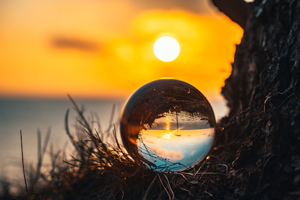 A crystal ball reflects a sunset over a calm body of water, with the sun low on the horizon. The colors of the sunset, including vibrant oranges and yellows, dominate the sky. The crystal ball is positioned on grass and dirt, adding texture to the foreground.