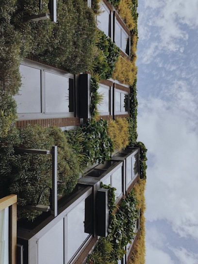 A vertical garden covers the facade of a building with lush greenery and various plant species arranged in a pattern alongside window panels. The sky is visible, featuring scattered clouds.
