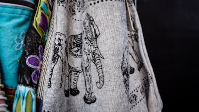 A detailed textile features intricate black elephant designs on a beige fabric. The fabric is part of a colorful arrangement including vibrant turquoise and patterns of purple flowers. The contrast between the darker designs and light fabric highlights the texture.