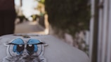 A sticker featuring blue cartoon eyes with expressive black eyebrows is displayed on a rounded, possibly concrete surface in an outdoor setting. In the blurry background, a pathway lined with flora and structures can be seen, indicating an urban or suburban environment.