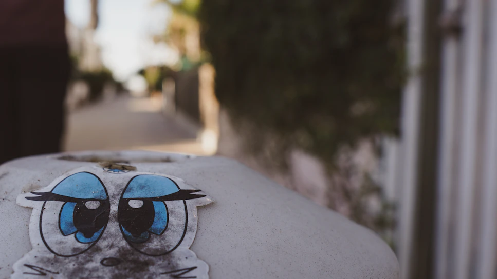 A sticker featuring blue cartoon eyes with expressive black eyebrows is displayed on a rounded, possibly concrete surface in an outdoor setting. In the blurry background, a pathway lined with flora and structures can be seen, indicating an urban or suburban environment.