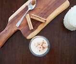 A wooden cutting board with a spoon containing ground cinnamon and several cinnamon sticks, accompanied by a glass of a creamy drink topped with sprinkled cinnamon.