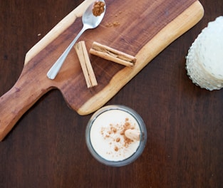 A wooden cutting board with a spoon containing ground cinnamon and several cinnamon sticks, accompanied by a glass of a creamy drink topped with sprinkled cinnamon.