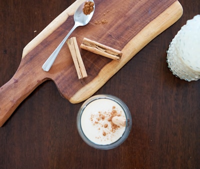 A wooden cutting board with a spoon containing ground cinnamon and several cinnamon sticks, accompanied by a glass of a creamy drink topped with sprinkled cinnamon.