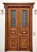 A large wooden double door with intricate carvings and metal grilles in the upper panels. The door features two ornate circular handles and is framed by a plain white wall. Two wall-mounted outdoor lights are placed symmetrically on either side of the door.
