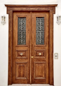 A large wooden double door with intricate carvings and metal grilles in the upper panels. The door features two ornate circular handles and is framed by a plain white wall. Two wall-mounted outdoor lights are placed symmetrically on either side of the door.